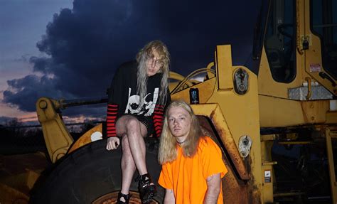 Laura Les and Dylan Brady, the two members of 100 gecs. Laura is sitting atop an excavator, with a can of Monster Energy next to her. Dylan is standing on the ground next to Laura.